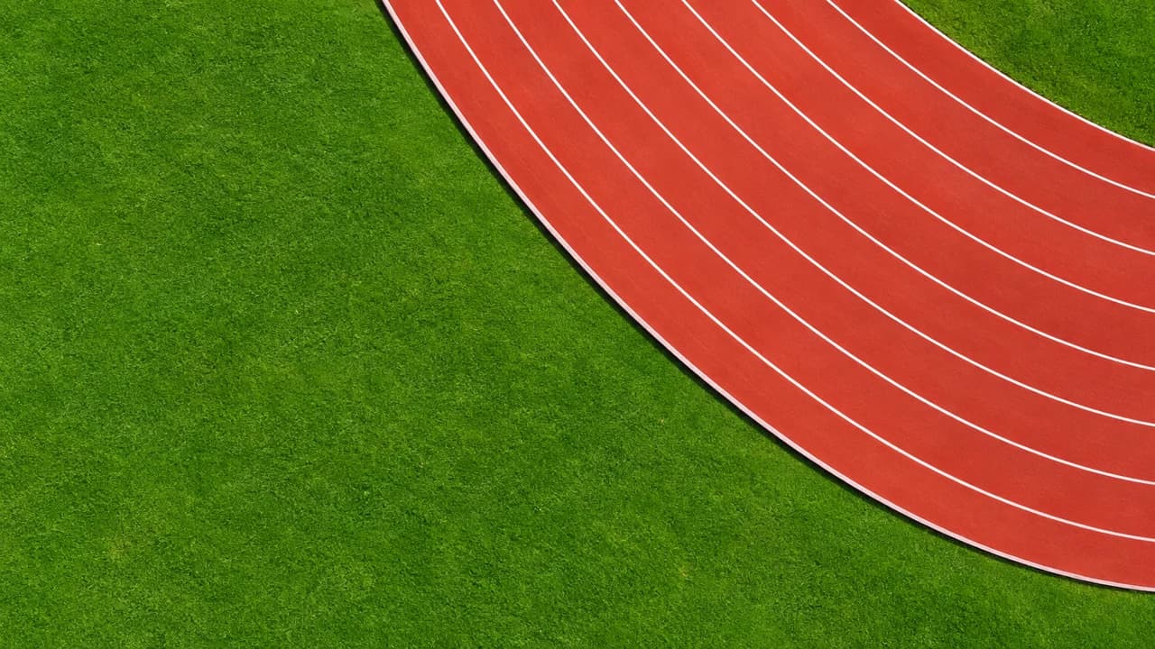 Top-down view of red running track lanes curving across the top right in hard midday light, plain green infield on the left.